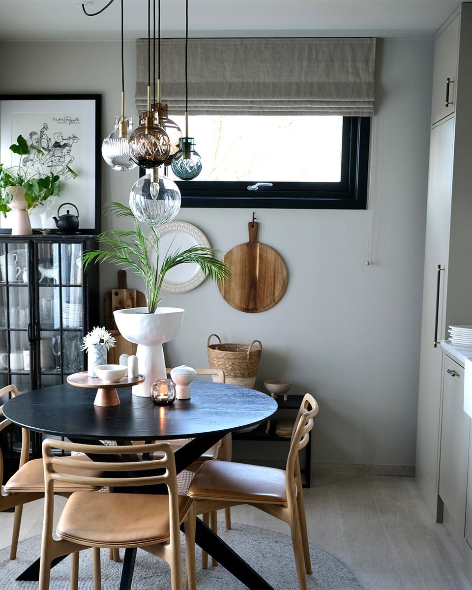 Lyst kjøkken med vindu med beige liftgardin i lin, rundt kjøkkenbord med stoler, vitrineskap, taklamper i glass og diverse pyntegjenstander. Foto.